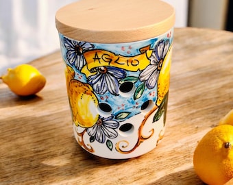 Peut inclure: Un pot à ail cylindrique en céramique avec un couvercle en bois clair, orné de citrons jaunes peints à la main, de fleurs bleues et d'une bannière jaune portant l'inscription AGLIO. Le récipient blanc présente plusieurs trous d'aération.