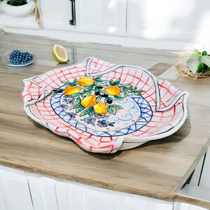 May include: A white ceramic serving platter with a red and blue checkered pattern and a hand-painted design of lemons and olive branches. The platter has a scalloped edge and is perfect for serving appetizers, snacks, or desserts.