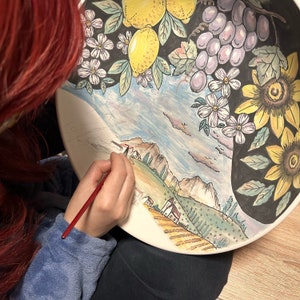 May include: A person paints a ceramic plate with a brush. The plate features a colorful landscape scene with mountains, houses, and a field of flowers. The top of the plate has a black and white design with lemons, grapes, and sunflowers.
