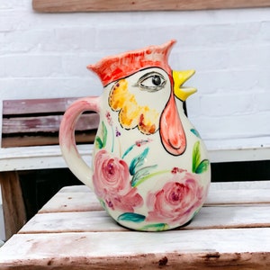 Könnte beinhalten: Eine weiße Keramikkanne in Form eines Hahns mit rotem Kamm, gelbem Schnabel und rosa und grünen Blumenmustern.