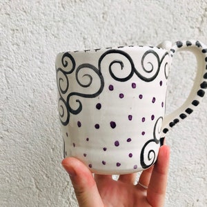 Pode incluir: Caneca de cer&acirc;mica branca com um design pintado &agrave; m&atilde;o em preto e roxo. O design apresenta redemoinhos e pontos.