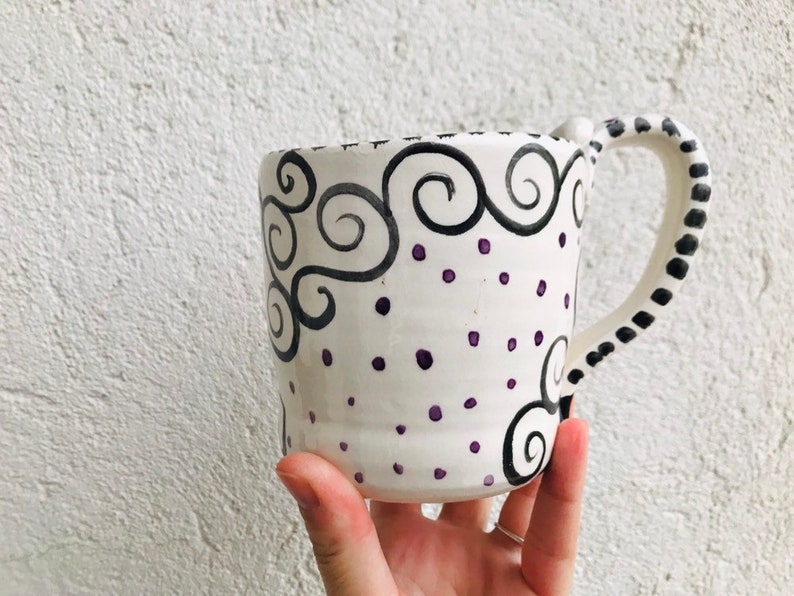 Pode incluir: Caneca de cer&acirc;mica branca com um design pintado &agrave; m&atilde;o em preto e roxo. O design apresenta redemoinhos e pontos.