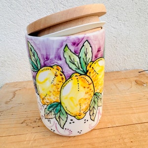 Puede incluir: Un recipiente cil&iacute;ndrico de cer&aacute;mica con un dise&ntilde;o pintado a mano de limones amarillo brillante y hojas verdes sobre un fondo de color violeta. El recipiente tiene una base blanca con peque&ntilde;os puntos negros y una tapa redonda de madera clara.