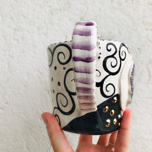 Pode incluir: Uma caneca de cer&acirc;mica branca com um design de redemoinhos preto e roxo. A al&ccedil;a &eacute; pintada com listras roxas. A caneca tem tachas douradas na parte inferior.