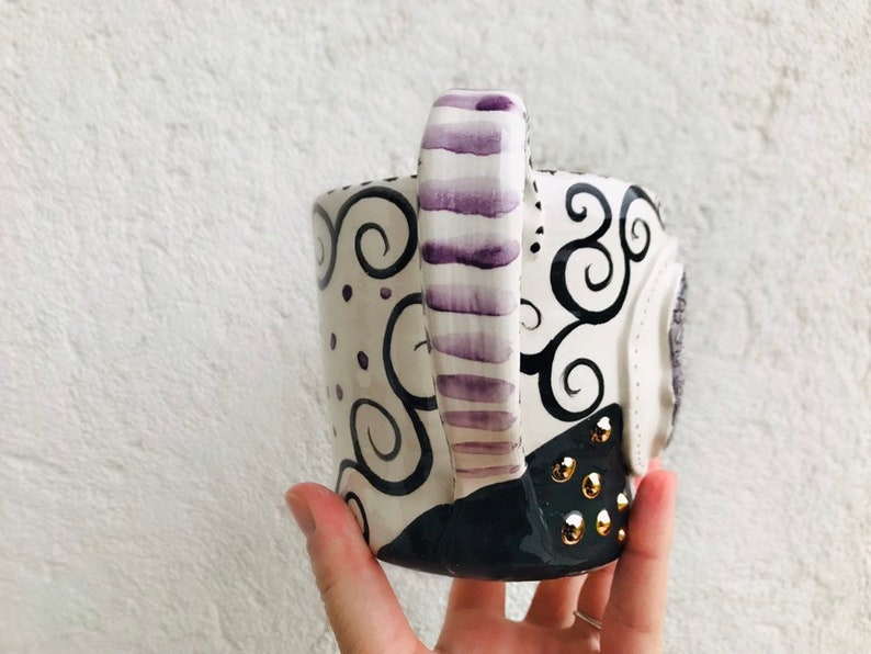 Pode incluir: Uma caneca de cer&acirc;mica branca com um design de redemoinhos preto e roxo. A al&ccedil;a &eacute; pintada com listras roxas. A caneca tem tachas douradas na parte inferior.