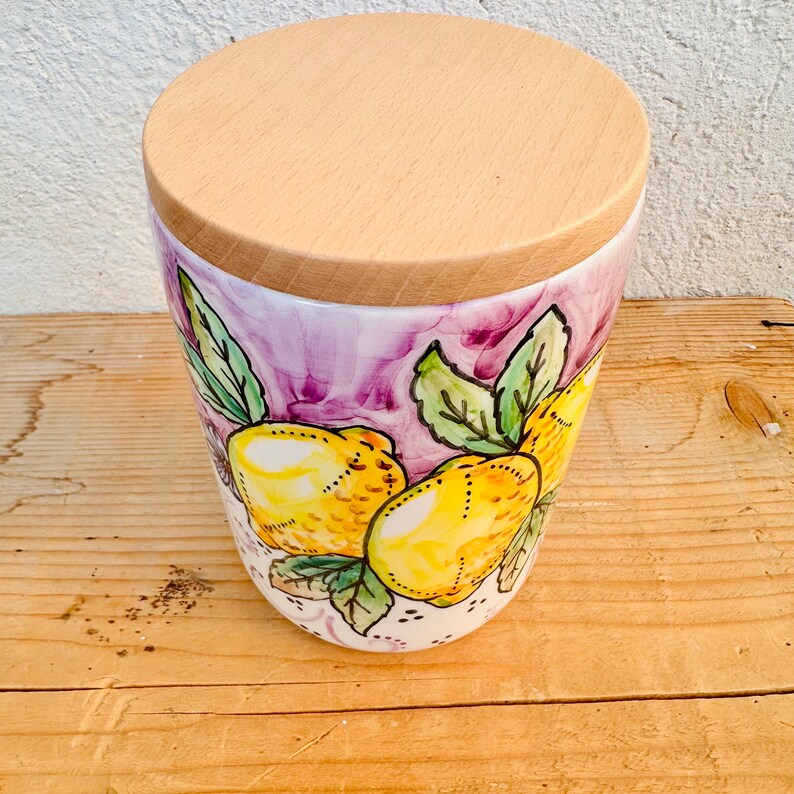 Puede incluir: Un recipiente cil&iacute;ndrico de cer&aacute;mica con un dise&ntilde;o pintado a mano de limones amarillos con hojas verdes sobre un fondo morado y blanco. El contenedor est&aacute; rematado con una tapa circular de madera clara que encaja al ras con el borde.