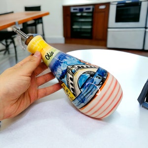 May include: A ceramic olive oil bottle with a cork stopper and a metal chain. The bottle is decorated with a colorful painting of a bridge and buildings. The bottle is labeled "Olio".