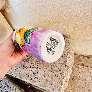 May include: A white ceramic vase with a purple and pink glaze. The vase is decorated with a hand-painted lemon design. The bottom of the vase has the text "Bottega Krua. Fatto a Mano in Italia."