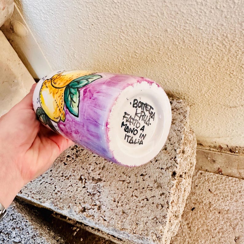 May include: A white ceramic vase with a purple and pink glaze. The vase is decorated with a hand-painted lemon design. The bottom of the vase has the text "Bottega Krua. Fatto a Mano in Italia."