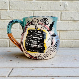 May include: A hand-painted ceramic mug with a whimsical design. The mug features a stack of books and mushrooms, with a blue handle and a light blue rim. The design includes black, white, yellow, pink, and orange accents.