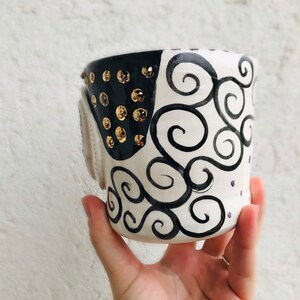 Pode incluir: Uma caneca de cer&acirc;mica branca com detalhes em preto e dourado. A caneca tem um design de espiral preto e branco e tachas douradas ao redor da borda.