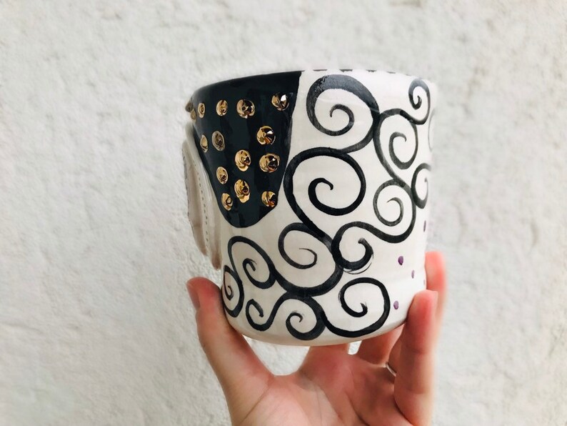 Pode incluir: Uma caneca de cer&acirc;mica branca com detalhes em preto e dourado. A caneca tem um design de espiral preto e branco e tachas douradas ao redor da borda.