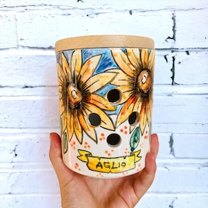 Könnte beinhalten: Eine weiße Keramikdose mit einem Holzdeckel, verziert mit Sonnenblumen und dem Wort "AGLIO" in einem Banner. Die Dose hat Löcher zur Belüftung.