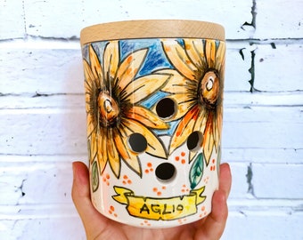 Peut inclure: Un pot en céramique blanche avec un couvercle en bois, décoré de tournesols et du mot "AGLIO" dans une bannière. Le pot est percé de trous pour la ventilation.