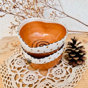 Peut inclure: Deux bols en céramique en forme de cœur avec une base blanche et un bord brun. Les bols sont décorés d'accents noirs et dorés. Le bol du haut a des pois blancs et un motif de spirale noire. Le bol du bas a un bord doré et un motif noir et blanc.