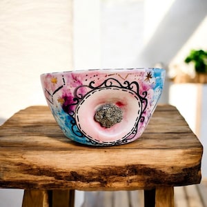 May include: A decorative ceramic bowl with a unique design. The bowl features a pink and blue watercolor background with black and gold accents. A central pink circle is framed by a black ornate design, and contains a cluster of metallic pyrite. The bowl sits on a wooden table.