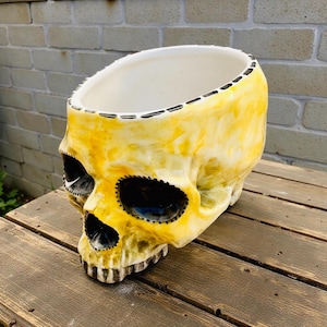 May include: A yellow skull-shaped ceramic planter with black eye sockets and a white interior. The planter has a stitched design around the rim and is placed on a wooden surface. The skull is painted with yellow and black hues.