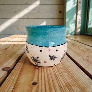 Puede incluir: Una maceta de cerámica hecha a mano con una parte superior azul turquesa y una base color crema. La base está decorada con ilustraciones de abejas negras y lunares rosas. La maceta está sobre una superficie de madera.