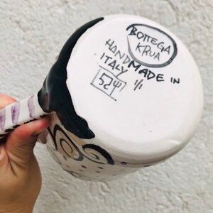 Pode incluir: Caneca de cer&acirc;mica branca com desenhos pintados em preto e roxo. A parte inferior da caneca tem o texto "HANDMADE IN ITALY" e "BOTTEGA KRUA" em um c&iacute;rculo.