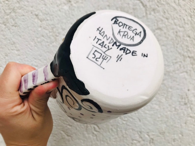 Pode incluir: Caneca de cer&acirc;mica branca com desenhos pintados em preto e roxo. A parte inferior da caneca tem o texto "HANDMADE IN ITALY" e "BOTTEGA KRUA" em um c&iacute;rculo.