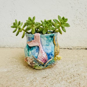 Op de afbeelding: Een keramische plantenpot met een gebeeldhouwde roze zeemeerminstaart en reliëfdetails van koraalrif in blauwe, groene en gouden tinten. De pot bevat een groene vetplant met dikke, puntige bladeren die uit de bovenste opening steken.