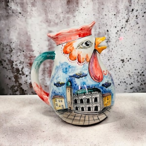 Puede incluir: Una jarra de cerámica blanca con forma de gallo con cresta y barbilla de color rojo, naranja y rosa. La jarra está decorada con un paisaje urbano azul y blanco y una asa verde.