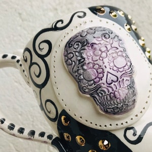 Pode incluir: Caneca de cer&acirc;mica branca com um design preto e branco que apresenta uma caveira de a&ccedil;&uacute;car com detalhes florais roxos e cinzentos. A caneca tem uma al&ccedil;a preta com bolinhas brancas e tachas douradas.