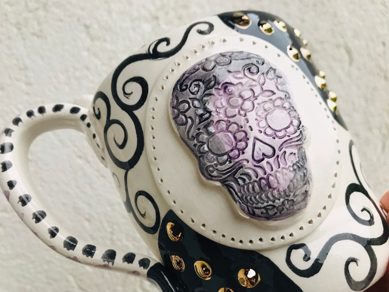 Pode incluir: Caneca de cer&acirc;mica branca com um design preto e branco que apresenta uma caveira de a&ccedil;&uacute;car com detalhes florais roxos e cinzentos. A caneca tem uma al&ccedil;a preta com bolinhas brancas e tachas douradas.
