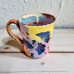 Op de afbeelding: Een handbeschilderde keramische mok met een kleurrijk ontwerp. De mok heeft een geel, paars en oranje kleurenschema met blauwe blad- en vlinderpatronen. De handgreep is oranje en bruin. De mok is ongeveer 10 cm hoog.