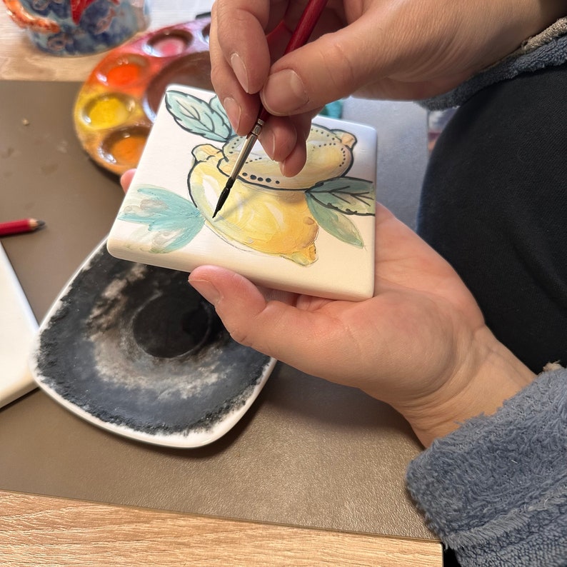 May include: A person paints a white ceramic tile with a black paintbrush. The tile features a hand-painted design of two lemons with green leaves.