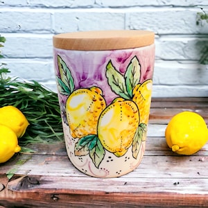 Puede incluir: Un recipiente cil&iacute;ndrico de cer&aacute;mica con tapa de madera clara, que presenta un dise&ntilde;o pintado a mano de limones amarillos brillantes con hojas verdes sobre un fondo crema y morado suave, acentuado con peque&ntilde;os puntos negros y espirales.