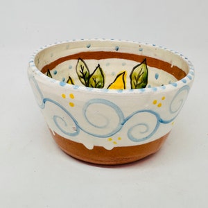 May include: A small, hand-painted ceramic bowl with a white glaze and brown rim. The bowl features a blue and white swirl design with yellow accents and green leaves.