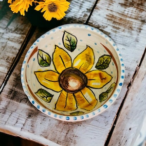 May include: A white ceramic bowl with a blue rim and a hand-painted sunflower design. The sunflower is yellow with a brown center and green leaves.