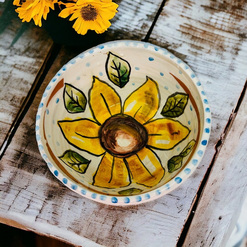 May include: A white ceramic bowl with a blue rim and a hand-painted sunflower design. The sunflower is yellow with a brown center and green leaves.