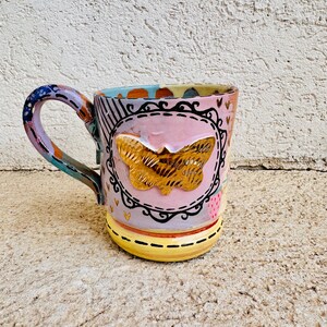 May include: A hand-painted ceramic mug with a butterfly design. The mug features a pink body with a gold butterfly in a black frame. The handle is multi-colored, and the base is yellow with black stripes. The mug is decorated with hearts and other patterns.