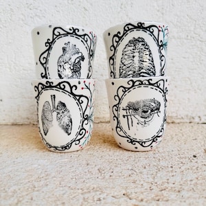 May include: Four white ceramic shot glasses with black anatomical illustrations of the heart, rib cage, lungs, and eye. Each glass has a black decorative border and small red and green accents.