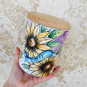 Puede incluir: Un tarro de cerámica blanco con tapa de madera, con un diseño pintado a mano de girasoles y la palabra "CAFFE" en azul. El tarro está decorado con detalles en morado, verde y amarillo.
