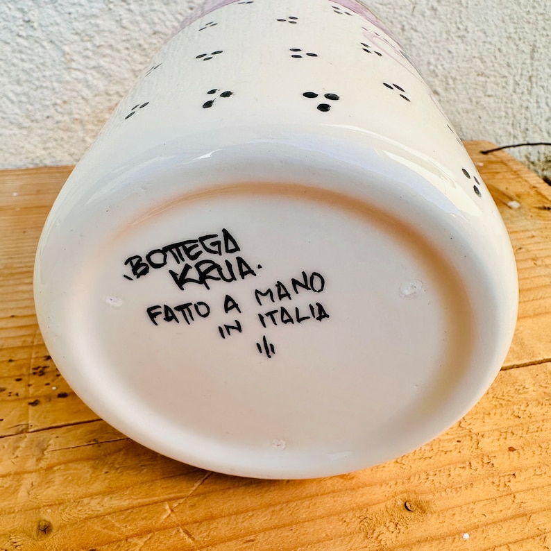 Puede incluir: Un recipiente de cer&aacute;mica blanca con un patr&oacute;n de peque&ntilde;os puntos negros. La base presenta texto negro escrito a mano que dice BOTTEGA KRUA, FATTO A MANO IN ITALIA y un peque&ntilde;o s&iacute;mbolo. La superficie es lisa, brillante y de forma cil&iacute;ndrica.