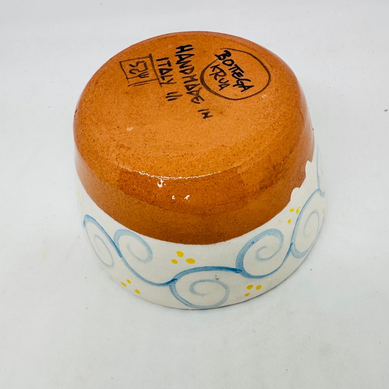 May include: A small, brown ceramic bowl with a white rim and blue and yellow swirl design. The bottom of the bowl is stamped with "HANDMADE IN ITALY" and "BOTTEGA KRUA".