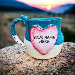May include: A blue and white ceramic mug with a heart-shaped design. The heart has the text "YOUR NAME HERE" written in black.