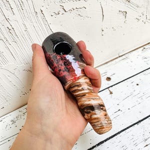 Könnte beinhalten: Eine handgehaltene Pfeife mit einer schwarzen Schale und einem gemusterten rot-weißen Design. Der Stiel hat ein braunes und hellbraunes marmoriertes Aussehen. Die Pfeife wird vor einem weißen Holzhintergrund gehalten.