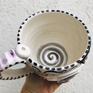 Pode incluir: Caneca de cer&acirc;mica branca com um design de espiral preto e branco no interior e uma al&ccedil;a listrada roxa e branca. A caneca tem uma borda preta e uma linha pontilhada preta ao redor da parte superior.