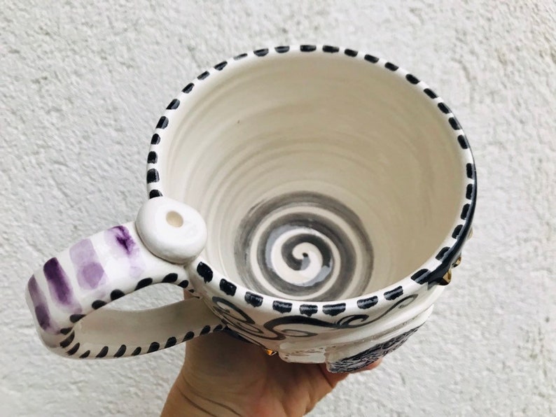 Pode incluir: Caneca de cer&acirc;mica branca com um design de espiral preto e branco no interior e uma al&ccedil;a listrada roxa e branca. A caneca tem uma borda preta e uma linha pontilhada preta ao redor da parte superior.