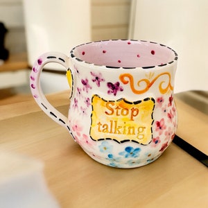 Puede incluir: Una taza de cerámica blanca con interior rosa y diseño floral. La taza tiene un asa negra y un letrero amarillo que dice "Stop talking".