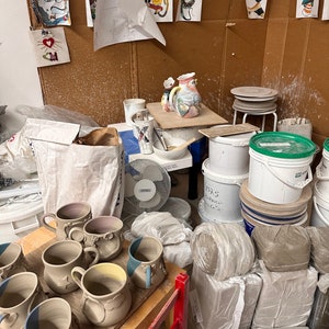 Puede incluir: Una colecci&oacute;n de tazas de cer&aacute;mica con cuerpos claros y bordes ba&ntilde;ados en azul, dispuestas sobre una superficie de madera. La escena incluye varios suministros de alfarer&iacute;a, cubos de pl&aacute;stico blanco, pilas de arcilla, un ventilador y dibujos.