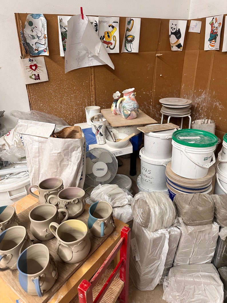 Puede incluir: Una colecci&oacute;n de tazas de cer&aacute;mica con cuerpos claros y bordes ba&ntilde;ados en azul, dispuestas sobre una superficie de madera. La escena incluye varios suministros de alfarer&iacute;a, cubos de pl&aacute;stico blanco, pilas de arcilla, un ventilador y dibujos.