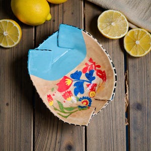 May include: Hand-painted ceramic bowl with a unique shape and floral design. The bowl features a light blue section and is decorated with red, blue, and yellow flowers. The rim is outlined with a black dotted pattern. Lemons are arranged around the bowl on a wooden surface.