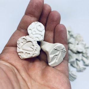 Może przedstawiać: Kolekcja małych, kremowych stempli ceramicznych z różnymi wytłoczonymi wzorami. Jeden stempel przedstawia serce, inny ma wzór wiru, a trzeci przedstawia motyw przypominający liść. Stemple są trzymane w dłoni.