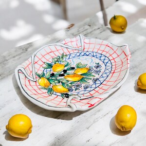 May include: A white ceramic serving dish with a red and blue checkered pattern and a lemon tree design. The dish has a ruffled edge and is surrounded by lemons.