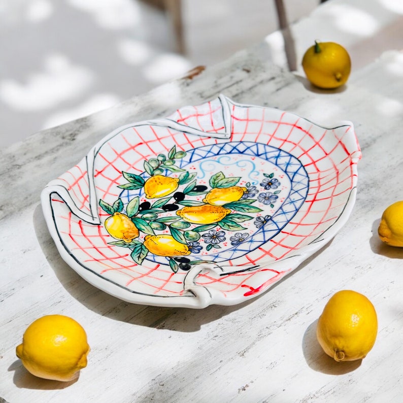 May include: A white ceramic serving dish with a red and blue checkered pattern and a lemon tree design. The dish has a ruffled edge and is surrounded by lemons.